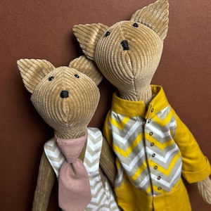 May include: Two plush toy foxes wearing different outfits. One fox is wearing a white and yellow chevron jumpsuit with a yellow collar. The other fox is wearing a white and yellow chevron dress with a pink tie.