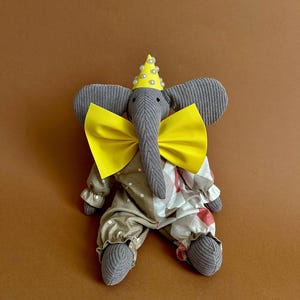 May include: A gray stuffed elephant wearing a yellow bow tie and a yellow party hat with white pearl accents. The elephant is sitting on a brown surface.