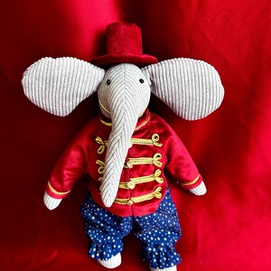 May include: A gray plush elephant wearing a red velvet jacket with gold trim, blue and gold star patterned pants, and a red top hat.