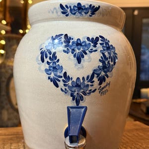 May include: A white ceramic pitcher with a blue and white floral design. The pitcher has a heart-shaped design with the word "Sandy" and the year "1992" written on it. The pitcher has a blue plastic spigot.