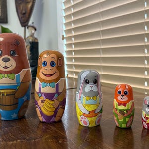May include: A set of six colorful wooden nesting dolls, or matryoshka dolls, featuring animal characters. The largest doll is a brown bear holding a honey pot, followed by a monkey holding bananas, a gray rabbit holding a carrot, a squirrel holding an acorn, and a gray mouse holding a piece of cheese.