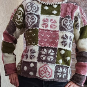 Op de afbeelding: Een patchwork trui met geometrische en bloemmotieven in wit, roze, groen en bruin. De trui heeft een ronde hals met een touwachtige rand en lange mouwen. Het ontwerp bestaat uit vierkanten met verschillende motieven.