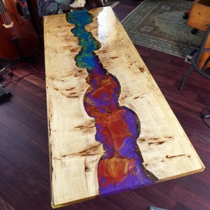 Peut inclure: Une table basse rectangulaire en bois avec une rivière de résine époxy colorée. Le bois a une finition claire et naturelle avec des motifs de grain sombres. La résine coule au centre en nuances de bleu, violet et orange.