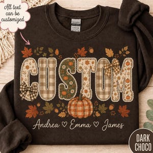 May include: A dark chocolate brown crewneck sweatshirt featuring the word CUSTOM in large, patterned letters with plaid and floral designs. The graphic includes autumn leaves, acorns, and a pumpkin, with the names Andrea, Emma, and James below.
