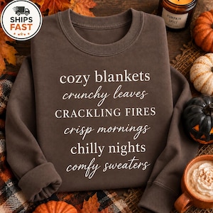 May include: A dark brown crewneck sweatshirt featuring white text that reads: cozy blankets, crunchy leaves, CRACKLING FIRES, crisp mornings, chilly nights, comfy sweaters. The garment has long sleeves and a ribbed collar, cuffs, and hemline.