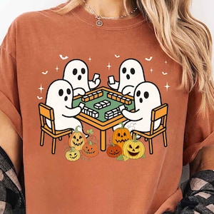 May include: A burnt orange t-shirt featuring a Halloween-themed graphic. The design shows four white ghost characters playing a game at a table, surrounded by jack-o'-lanterns. The ghosts are seated on yellow chairs, and bats are flying above.