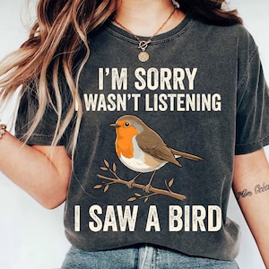 May include: Dark gray t-shirt with the text "I'M SORRY I WASN'T LISTENING I SAW A BIRD". The shirt features a graphic of a robin perched on a branch. The bird has orange, brown, and white coloring.