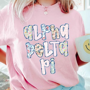 May include: A light pink t-shirt with the text "Alpha Delta Pi" in a floral and gingham pattern. A white mug with a yellow smiley face is held in the hand.
