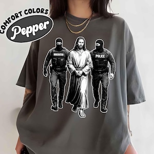 May include: Pepper Comfort Colors t-shirt featuring a graphic of Jesus being escorted by two figures labeled "Immigration" and "Police." The design is in black and white on a gray shirt. The shirt has short sleeves.