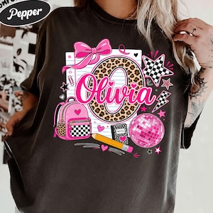 May include: A charcoal gray t-shirt featuring a graphic print with the name Olivia in pink script. The design includes a large leopard print letter O, a pink bow, a pink backpack, a yellow pencil, a disco ball, and a notebook labeled Notes.