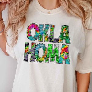 May include: White t-shirt with a colorful graphic design that says "OKLAHOMA" with a weather radar map pattern.
