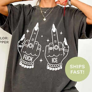 Puede incluir: Camiseta color pimienta con un gráfico blanco de dos manos mostrando el dedo medio. La palabra "FUCK" está en una mano y "ICE" en la otra. Las manos están adornadas con anillos y pulseras.