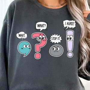 May include: Dark gray sweatshirt with cartoon speech bubbles and characters. The design includes a blue speech bubble with "WAIT," a pink question mark with eyes, a gray rock with "STOP IT," a white speech bubble with "WHAT?", and a purple exclamation point with "I AGREE!"