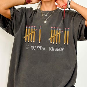 May include: Dark gray t-shirt with a graphic of pencils and the text "IF YOU KNOW... YOU KNOW". The pencils are yellow with pink erasers. The shirt is a casual, oversized style.