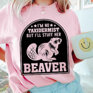 May include: A light pink t-shirt with a black and white graphic of a beaver and the text "I'M NO TAXIDERMIST BUT I'LL STUFF HER BEAVER". A white mug with a yellow smiley face is in the background.