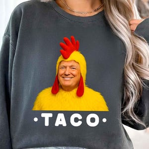 Puede incluir: Sudadera gris oscuro con una imagen humorística de una persona con un disfraz de pollo. El disfraz es amarillo con detalles rojos y una cara sonriente. La palabra "TACO" está impresa en blanco debajo.