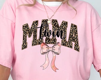 May include: Pink t-shirt with the word "MAMA" in leopard print block letters, with the word "twin" in cursive script in the middle. A pink bow with watercolor-style ribbons is below the text.