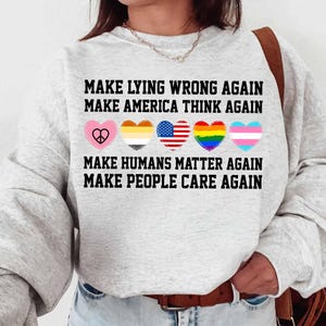 May include: A gray sweatshirt with the text "Make Lying Wrong Again, Make America Think Again, Make Humans Matter Again, Make People Care Again". The text is in black and there are five hearts with different symbols in them: a peace sign, a rainbow, a transgender symbol, an American flag, and a symbol for the LGBTQ+ community.