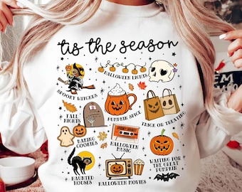 May include: White long-sleeve sweatshirt with a Halloween graphic. The design features a witch, pumpkin spice, a skull, and the text "tis the season". Other elements include a black cat, a tombstone, and a jack-o'-lantern.