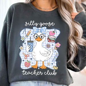 K&ouml;nnte beinhalten: Dunkelgraues Sweatshirt mit einer skurrilen Grafik einer Gans mit Brille und Schleife, die ein Getr&auml;nk h&auml;lt. Das Design enth&auml;lt den Text "silly goose teacher club" mit Illustrationen zum Thema Schule.