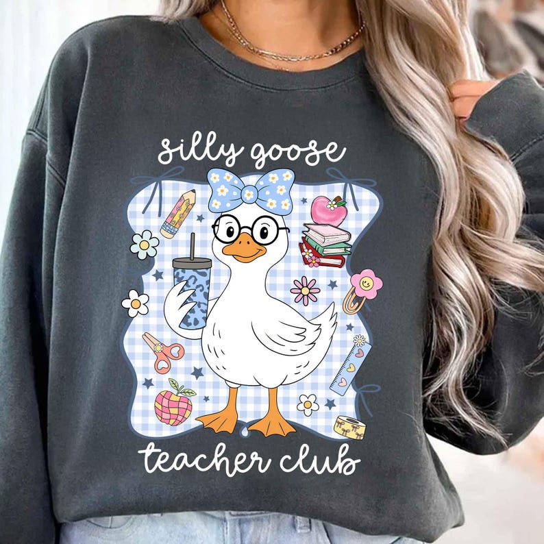 K&ouml;nnte beinhalten: Dunkelgraues Sweatshirt mit einer skurrilen Grafik einer Gans mit Brille und Schleife, die ein Getr&auml;nk h&auml;lt. Das Design enth&auml;lt den Text "silly goose teacher club" mit Illustrationen zum Thema Schule.