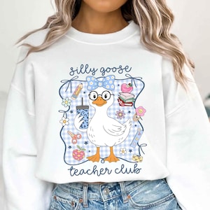 K&ouml;nnte beinhalten: Wei&szlig;es Sweatshirt mit einer Grafik einer Gans mit Brille und Schleife, die ein Getr&auml;nk h&auml;lt. Der Text "silly goose teacher club" ist zu sehen. Das Design enth&auml;lt schulthematische Elemente wie Bleistifte, Blumen und einen Apfel.