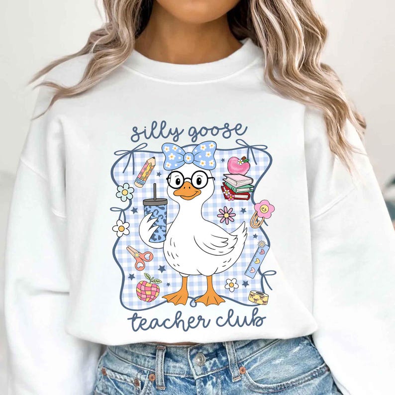 K&ouml;nnte beinhalten: Wei&szlig;es Sweatshirt mit einer Grafik einer Gans mit Brille und Schleife, die ein Getr&auml;nk h&auml;lt. Der Text "silly goose teacher club" ist zu sehen. Das Design enth&auml;lt schulthematische Elemente wie Bleistifte, Blumen und einen Apfel.
