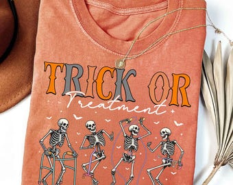 May include: A coral-colored t-shirt with the words "TRICK OR TREATMENT" in orange and gray. The shirt features a graphic of dancing skeletons with various props, including a walker and jump rope. A gold necklace is draped across the shirt.