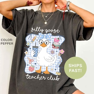 K&ouml;nnte beinhalten: Pfefferfarbenes T-Shirt mit einem Cartoon-Gans-Design, das eine Brille und eine Schleife tr&auml;gt und ein Getr&auml;nk h&auml;lt. Der Text lautet "silly goose teacher club" mit Illustrationen zum Thema Schule. Das Shirt hat eine lockere Passform.