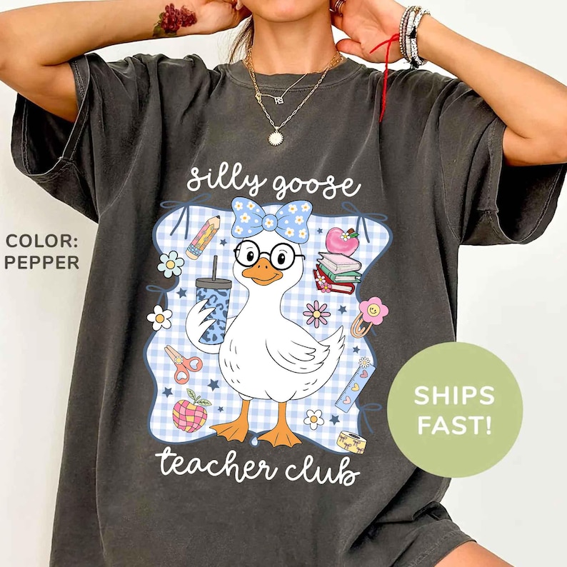K&ouml;nnte beinhalten: Pfefferfarbenes T-Shirt mit einem Cartoon-Gans-Design, das eine Brille und eine Schleife tr&auml;gt und ein Getr&auml;nk h&auml;lt. Der Text lautet "silly goose teacher club" mit Illustrationen zum Thema Schule. Das Shirt hat eine lockere Passform.