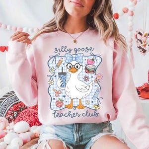 K&ouml;nnte beinhalten: Hellrosa Sweatshirt mit einer Cartoon-Gans, die eine Brille und eine Schleife tr&auml;gt und ein Getr&auml;nk h&auml;lt. Das Design enth&auml;lt den Text "silly goose teacher club" und schulische Grafiken wie Bleistifte und B&uuml;cher.