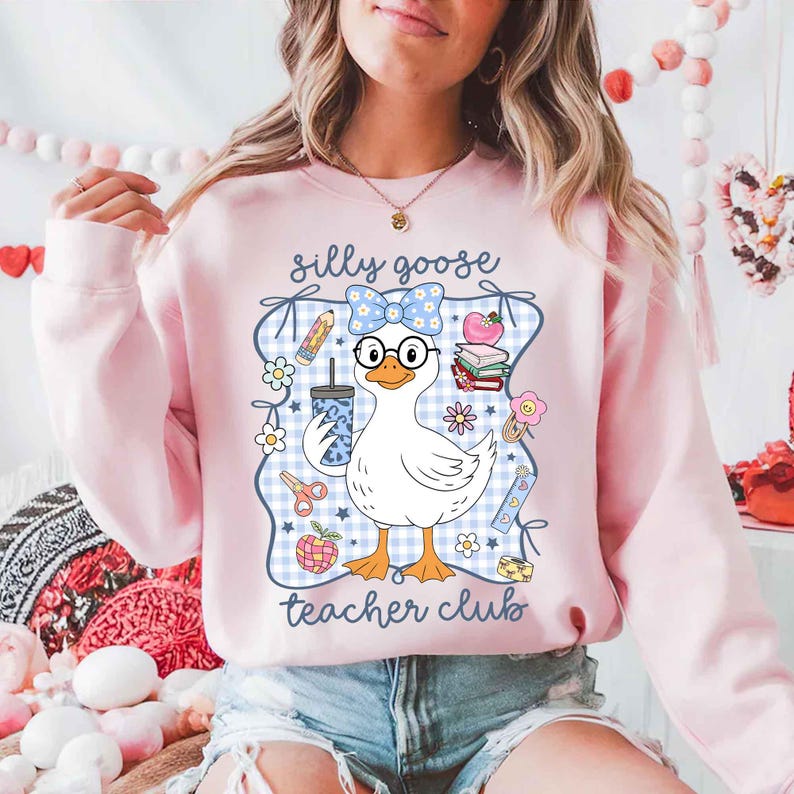 K&ouml;nnte beinhalten: Hellrosa Sweatshirt mit einer Cartoon-Gans, die eine Brille und eine Schleife tr&auml;gt und ein Getr&auml;nk h&auml;lt. Das Design enth&auml;lt den Text "silly goose teacher club" und schulische Grafiken wie Bleistifte und B&uuml;cher.