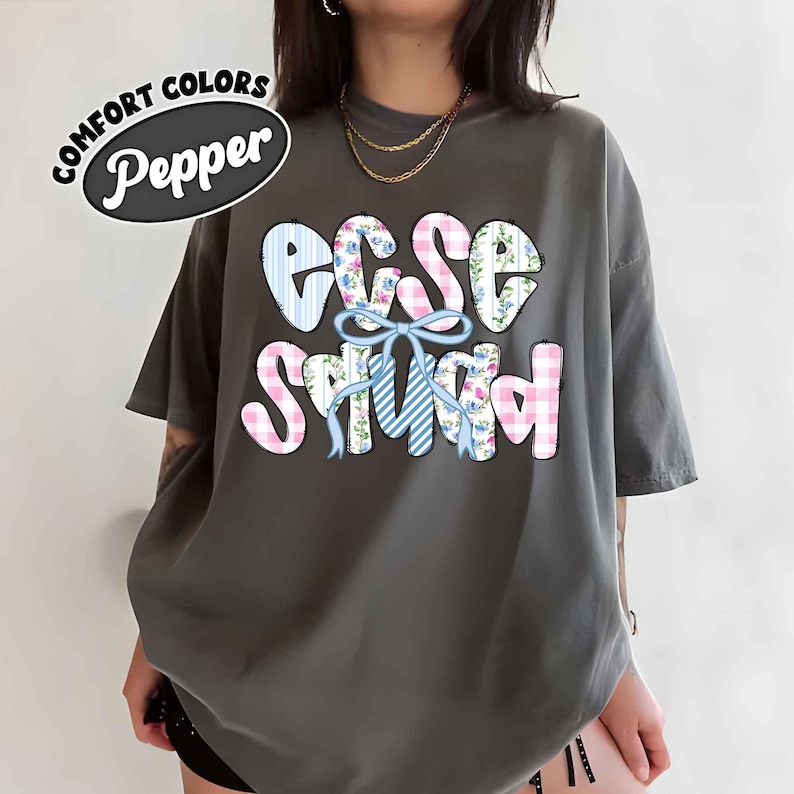 Puede incluir: Una camiseta Comfort Colors gris oscuro con el texto "ECCE Squad" en una fuente l&uacute;dica. Las letras est&aacute;n decoradas con patrones de cuadros azules y rosas, estampados florales y un lazo azul. La palabra "Pepper" est&aacute; en un dise&ntilde;o circular.