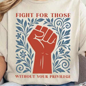 May include: Cream-colored t-shirt featuring a red fist graphic, surrounded by blue floral and leaf designs. The text "FIGHT FOR THOSE WITHOUT YOUR PRIVILEGE" is printed in red. The shirt has short sleeves.