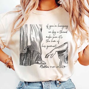 May include: A white t-shirt featuring a black and white illustration of a person reaching out their hand. The text on the shirt reads "if you're hanging on by a thread make sure it's the hem of his garment. Matthew 9:20-27".
