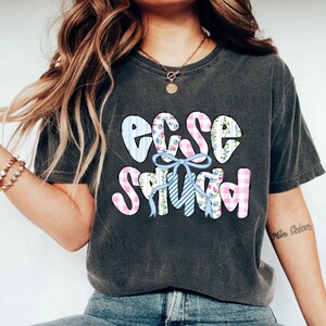 Puede incluir: Camiseta gris oscuro con el texto "EGG SQUAD" en un dise&ntilde;o de patchwork colorido. Las letras est&aacute;n decoradas con cuadros vichy azules y rosas, estampados florales y un lazo azul.