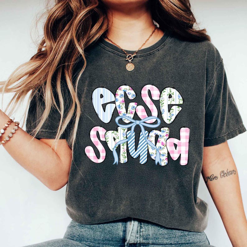Puede incluir: Camiseta gris oscuro con el texto "EGG SQUAD" en un dise&ntilde;o de patchwork colorido. Las letras est&aacute;n decoradas con cuadros vichy azules y rosas, estampados florales y un lazo azul.