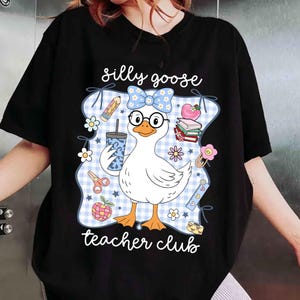 K&ouml;nnte beinhalten: Schwarzes T-Shirt mit einer Cartoon-Gans, die eine Brille und eine Schleife tr&auml;gt und ein Getr&auml;nk h&auml;lt. Das Design enth&auml;lt den Text "silly goose teacher club" mit Illustrationen zum Thema Schule.