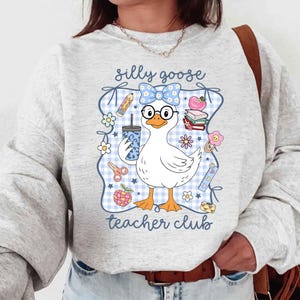 K&ouml;nnte beinhalten: Hellgraues Sweatshirt mit einem Cartoon-Gans-Design. Die Gans tr&auml;gt eine Brille und eine Schleife und h&auml;lt ein Getr&auml;nk. Das Design enth&auml;lt den Text "silly goose teacher club" mit schulischen Grafiken wie Bleistiften, B&uuml;chern und Blumen.