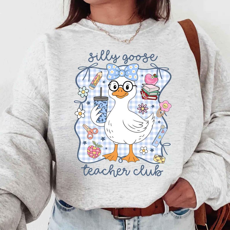 K&ouml;nnte beinhalten: Hellgraues Sweatshirt mit einem Cartoon-Gans-Design. Die Gans tr&auml;gt eine Brille und eine Schleife und h&auml;lt ein Getr&auml;nk. Das Design enth&auml;lt den Text "silly goose teacher club" mit schulischen Grafiken wie Bleistiften, B&uuml;chern und Blumen.