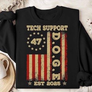 May include: Black sweatshirt with a distressed American flag design. The flag features red and beige stripes, with a circle of stars and the number "47". The text "TECH SUPPORT D.O.G.E. EST 2025" is printed above and below the flag.