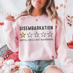 Op de afbeelding: Een lichtroze sweatshirt met het woord "DISEMBARKATION" in zwarte blokletters boven een beoordeling van één gouden ster en vier omrande sterren. De tekst eronder luidt "AWFUL, WOULD NOT RECOMMEND."