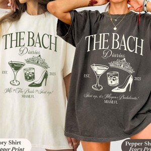 May include: Two oversized t-shirts, one ivory and one pepper, feature a graphic design with the text "THE BACH Diaries" and illustrations of a cocktail, high heel, and crown. The shirts also include the text "EST. 2025" and "MIAMI FL".