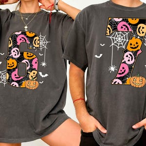May include: Two dark gray t-shirts with large, colorful Halloween-themed numbers 6 and 7. The numbers are filled with jack-o'-lanterns, spiderwebs, and bats. A pumpkin is at the base of each number.