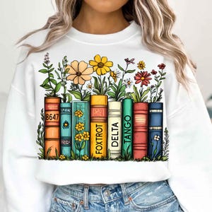 Op de afbeelding: Wit sweatshirt met een kleurrijke afbeelding van een rij boeken met bloemen die eruit groeien. De boeken hebben de woorden "Tango", "Delta", "Foxtrot" en "8647" op hun ruggen.