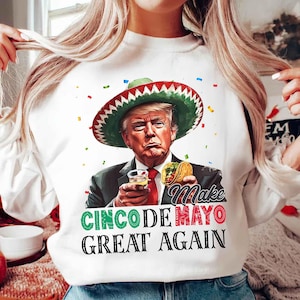 Peut inclure: T-shirt blanc avec un imprimé de Donald Trump portant un sombrero, tenant une margarita et un taco. Le texte "Make Cinco De Mayo Great Again" est imprimé sur le t-shirt.