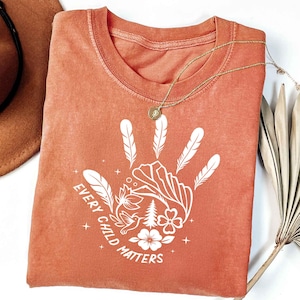 May include: Rust-colored t-shirt with a white handprint design featuring feathers, flowers, and the text "EVERY CHILD MATTERS." A gold necklace with a pendant rests on the shirt. The shirt is folded, and a brown hat is visible.