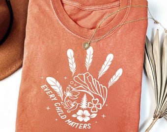 May include: A terracotta-coloured t-shirt with a white handprint design incorporating feathers, flowers, and the text "EVERY CHILD MATTERS." A gold necklace with a pendant lies on the shirt. The shirt is folded, and a brown hat is visible.
