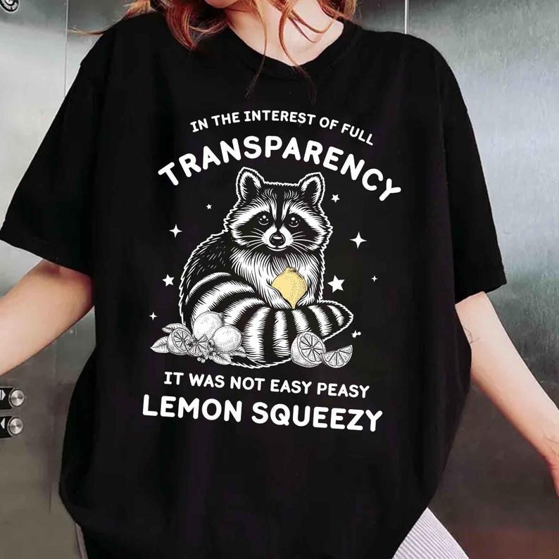 May include: Black t-shirt with a raccoon graphic holding a lemon, surrounded by text that reads "IN THE INTEREST OF FULL TRANSPARENCY" and "IT WAS NOT EASY PEASY LEMON SQUEEZY". The design includes stars and lemon slices.