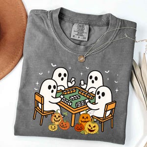 May include: Gray Comfort Colors t-shirt featuring a Halloween-themed graphic. The design shows four cartoon ghosts playing a game at a table, surrounded by pumpkins. A silver necklace with a pendant is draped across the shirt.